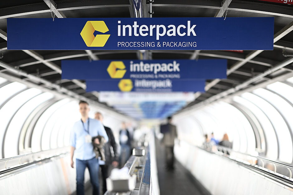 People walking along a conveyor belt beneath several blue signs featuring the yellow interpack logo and the text “interpack PROCESSING & PACKAGING” in a tunnel-like structure.